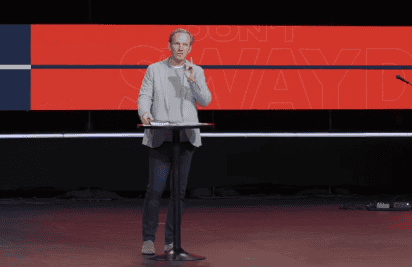 Pastor Steve Robinson on stage behind a podium, speaking with one hand raised. A large red and black screen with faint text is displayed in the background.