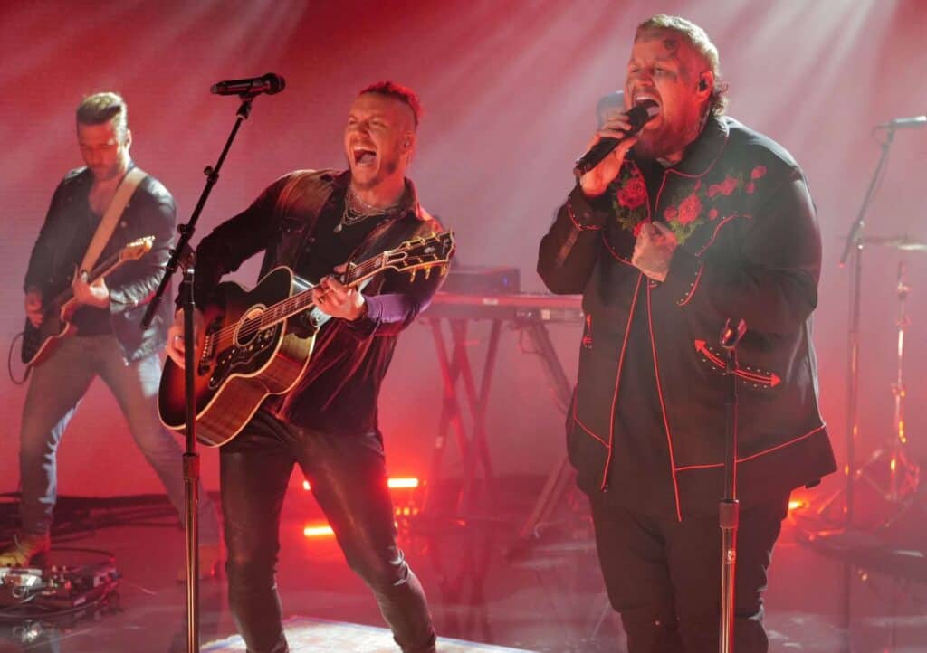 Four male musicians perform energetically on stage with red lighting; two play guitars, and one passionately sings into a microphone, weaving music storytelling into their set while wearing a black jacket with red accents.