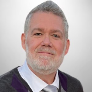 A middle-aged man with gray hair and beard wearing a gray sweater, white shirt, and silver tie, posing against a plain light background.