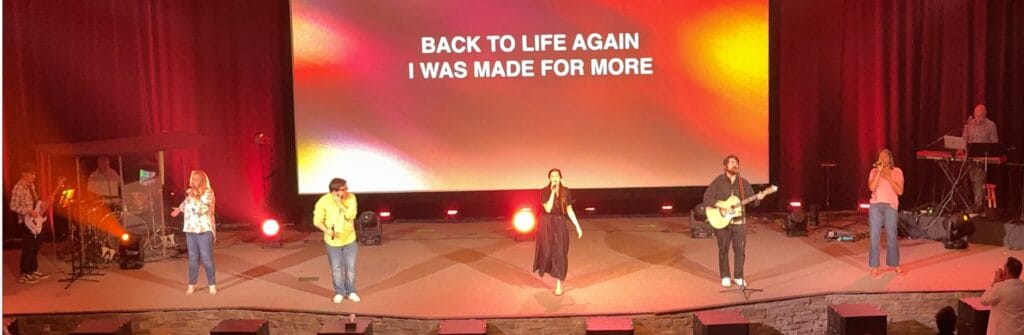 Six musicians stand on a stage under colorful lighting, performing with instruments. Behind them, a large screen displays the lyrics "BACK TO LIFE AGAIN I WAS MADE FOR MORE," inspiring the crowd with hope and purpose.