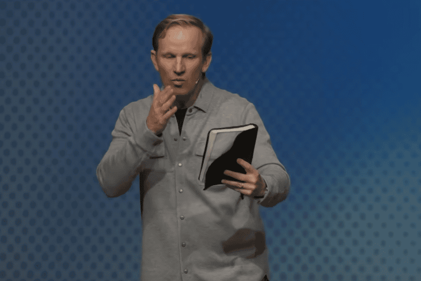 A man in a gray button-up shirt stands against a blue, dotted background. He holds an open notebook in one hand and gestures with his other, as if speaking words of inspiration or explaining hope through tribulation.