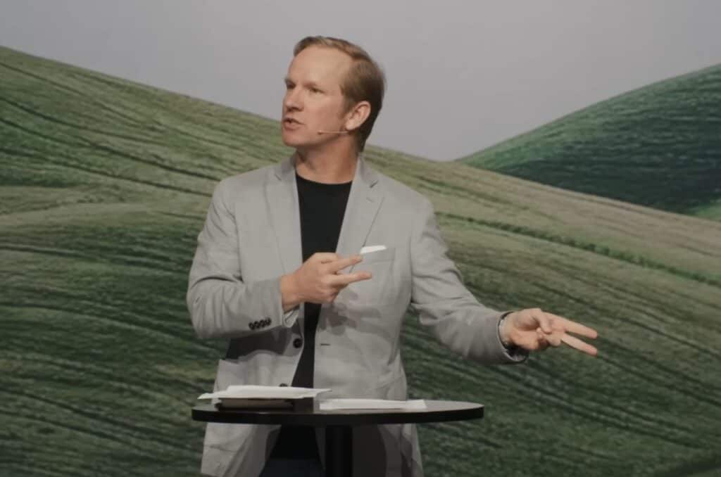 A man in a light gray blazer speaks into a headset microphone while gesturing with his hands. He stands behind a small round table with papers on it, with a backdrop of green rolling hills.