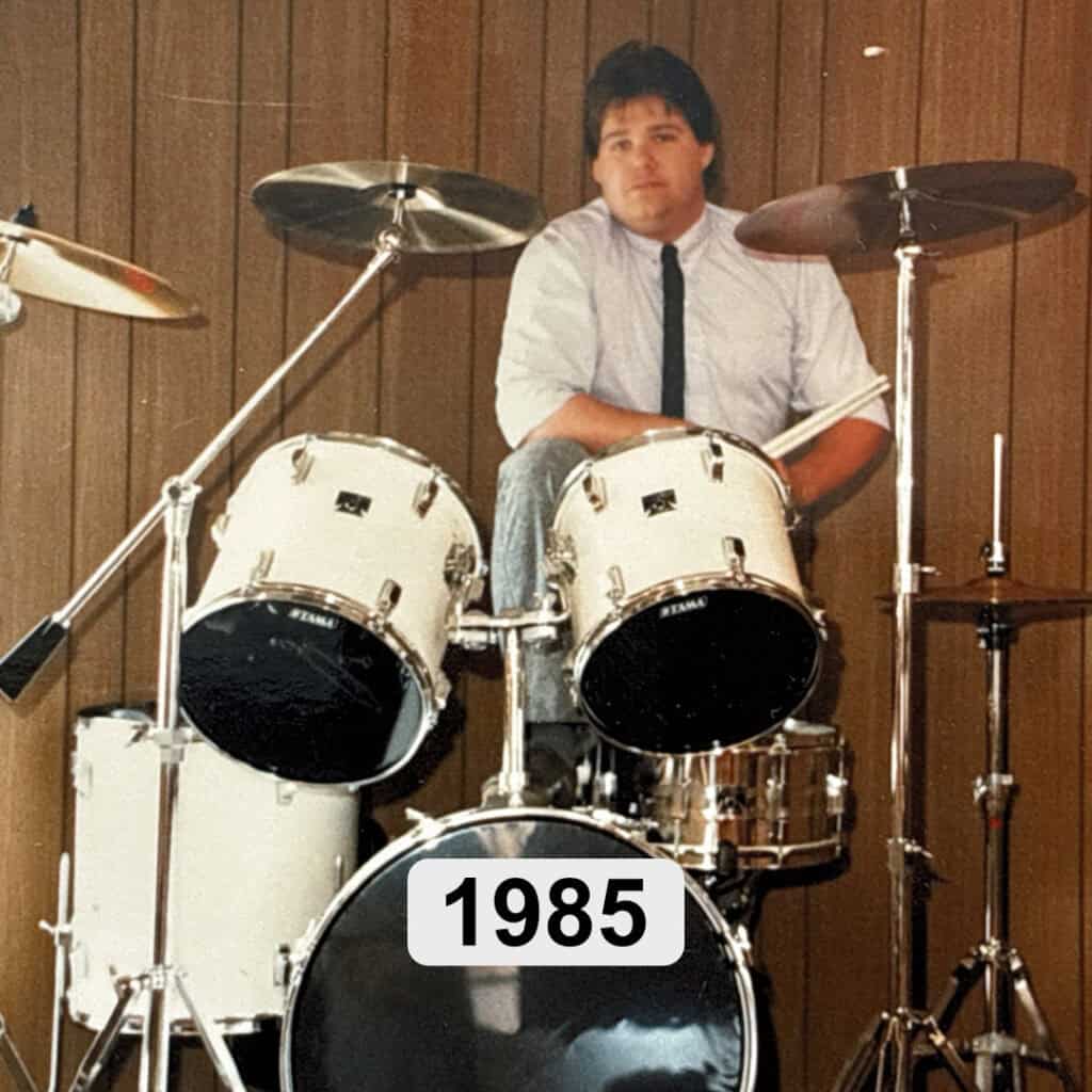 Kelly, in a white shirt and black tie sits at a white drum set in front of wood paneling. The drums are labeled "1985," hinting at a personal salvation testimony captured that year.