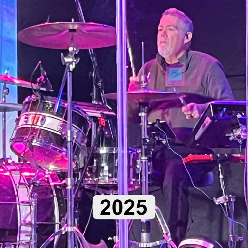 Kelly plays a drum set behind a clear barrier on a stage with purple lighting, appearing focused in his gray shirt. The number "2025" is displayed prominently, suggesting a powerful christian testimony unfolding through music.