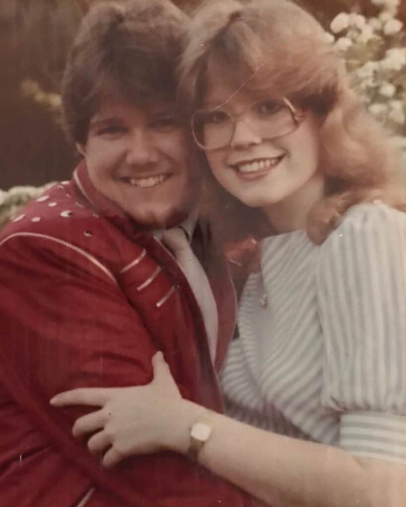 Kelly and Nancy smiles and embrace, radiating joy like a heartfelt christian testimony. Kelly wears a red jacket with white stripes; Nancy, in glasses and a striped dress. Flowers and greenery are blurred in the background.