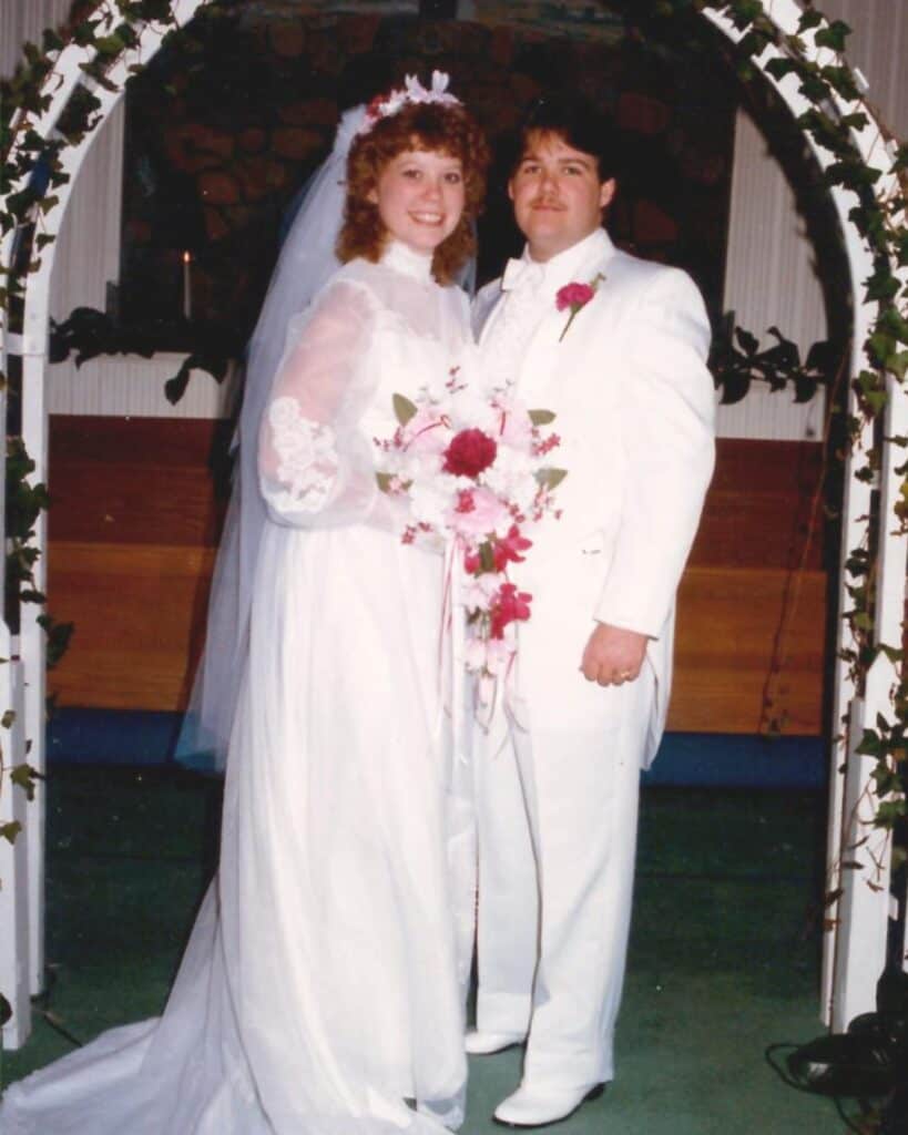 Nancy and Kelly stand together under a flower-decorated arch at their wedding, celebrating not only their love but also sharing how Jesus changed my life as part of their faith journey testimony.