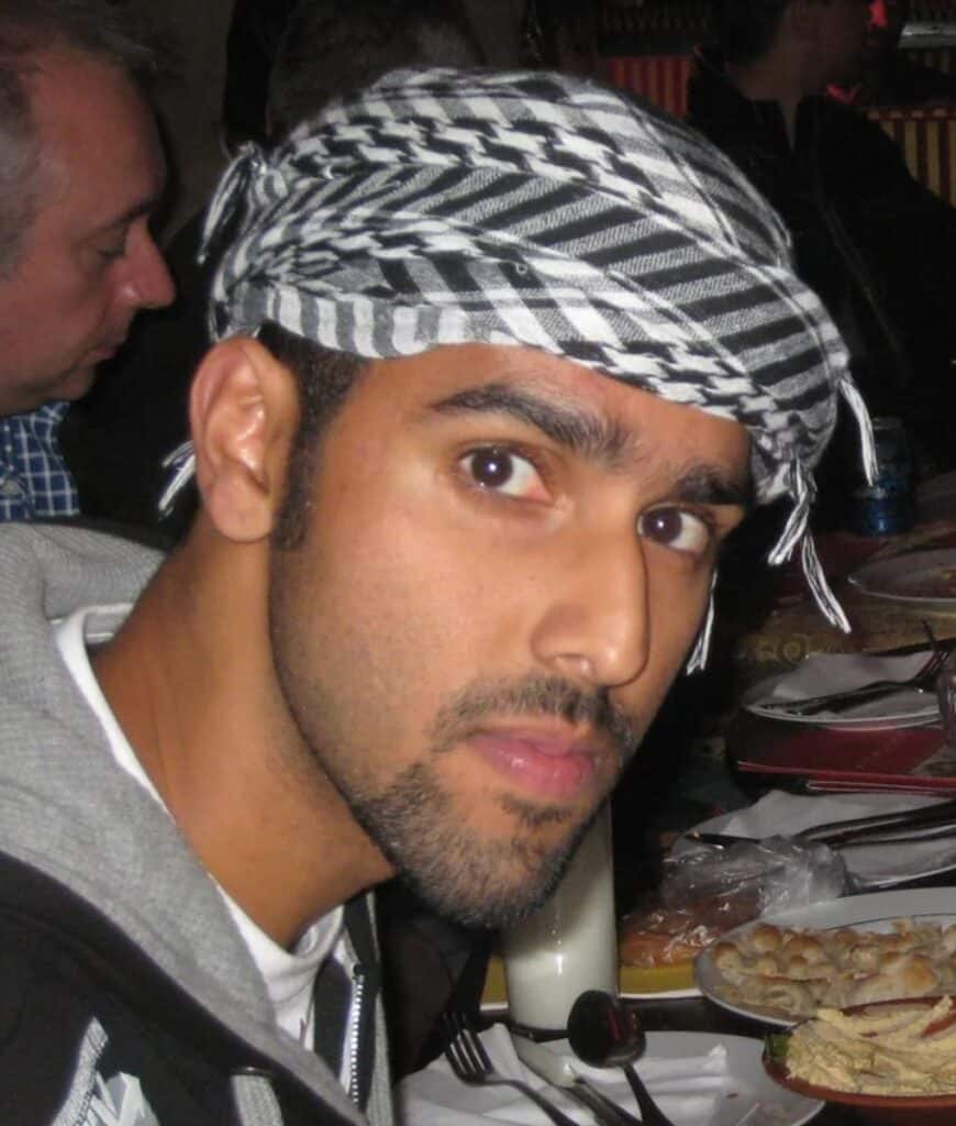 A man with a neatly trimmed beard and mustache wears a patterned headscarf and a dark jacket, looking toward the camera while seated at a table with plates of food—perhaps sharing thoughts on Nabeel Qureshi's "Seeking Allah, Finding Jesus." Other people dine in the background.
